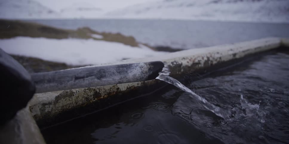 Crystal clear water flows from an old pipe into a trough on a cold, snowy day in a remote, wintery landscape in slow motion