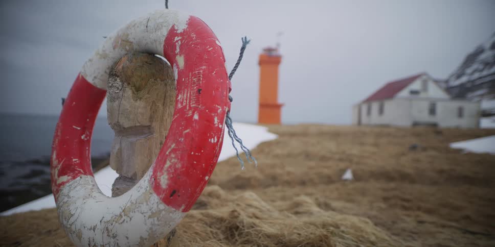 A weathered life preserver hangs on a carved wooden post with a bright orange lighthouse in the foggy background of a desolate Icelandic coast