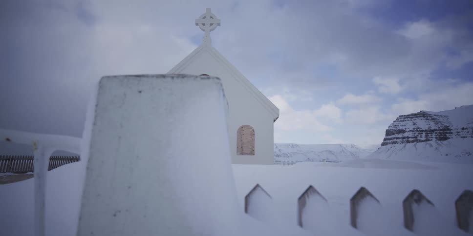 A slow tracking shot passes a snow-covered fence revealing a quaint white church with a red door set against a dramatic winter mountainscape