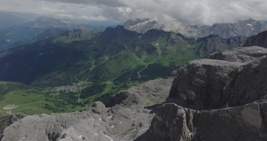 Drone footage slowly glides over a rocky mountain pass, revealing a stunning green valley and distant snow-capped peaks in the Italian Dolomites