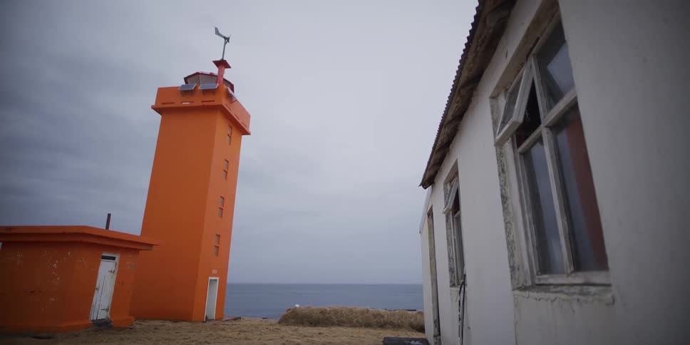 A stark orange lighthouse stands beside a derelict building overlooking the sea on a bleak, overcast day