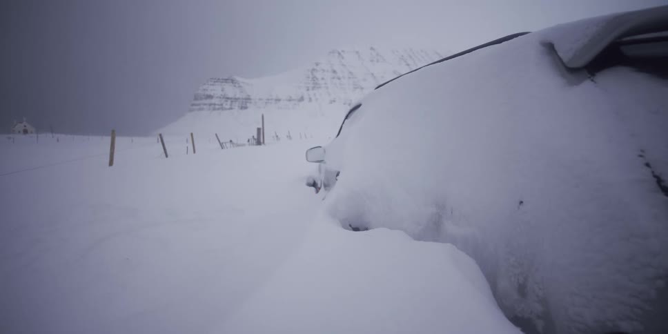 A car is almost completely buried in deep snow in a remote, mountainous landscape during a winter storm