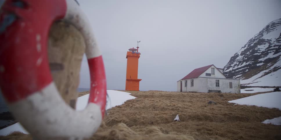 A striking orange lighthouse and an old house stand on a desolate, snowy coastline in Iceland, with a life preserver in the foreground