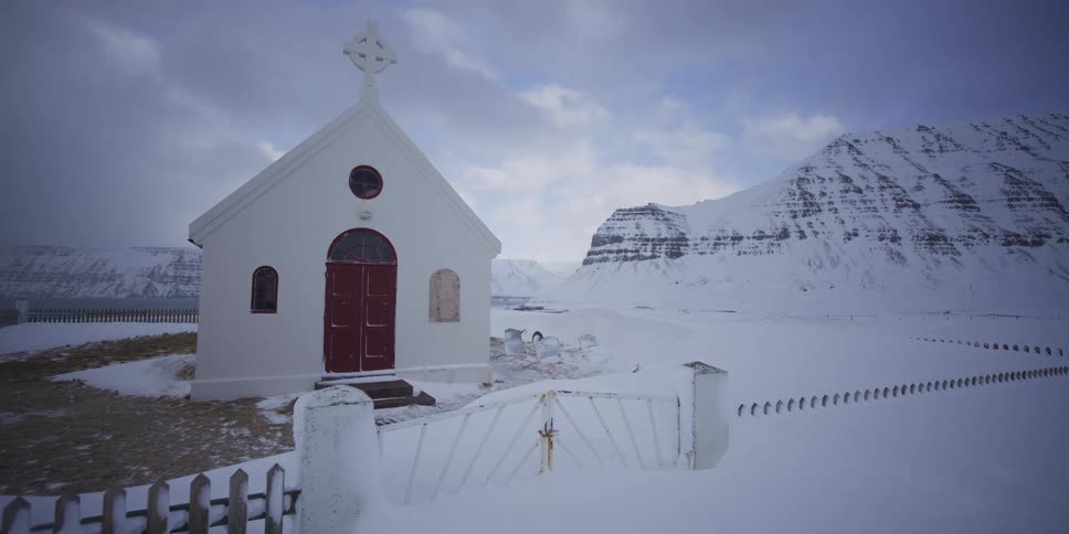 A small white church with a red door stands in a vast, snow-covered landscape at the base of a mountain range