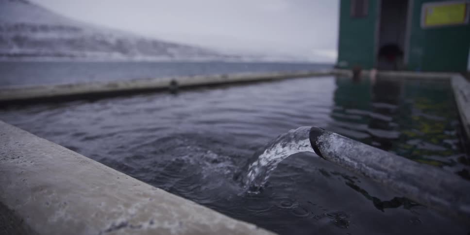 A serene slow-motion close-up shows a pipe pouring hot thermal water into an outdoor pool with a snowy Icelandic fjord in the background