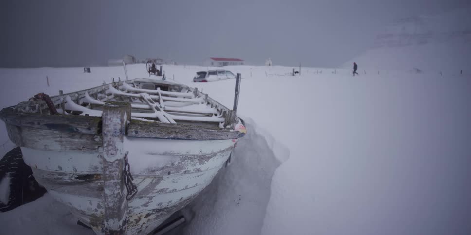 A slow dolly shot reveals an abandoned wooden boat trapped in deep snow during a harsh winter storm in a remote village