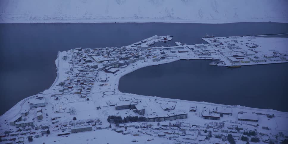 An aerial shot captures a quiet, snow-covered coastal town and its harbor nestled in a fjord during the blue hour of a winter day