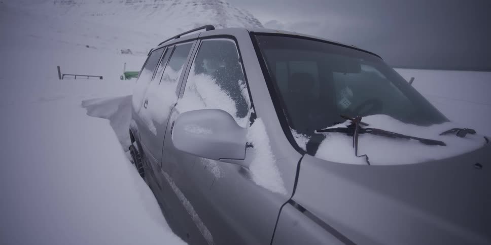 A silver SUV is almost completely buried in a deep snowdrift in a remote, mountainous winter landscape