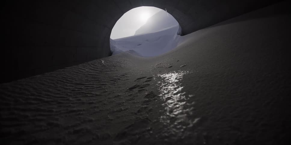 A slow pull-back shot from inside a dark, sandy culvert reveals water trickling towards a bright, snow-covered mountain landscape at the end of the tunnel