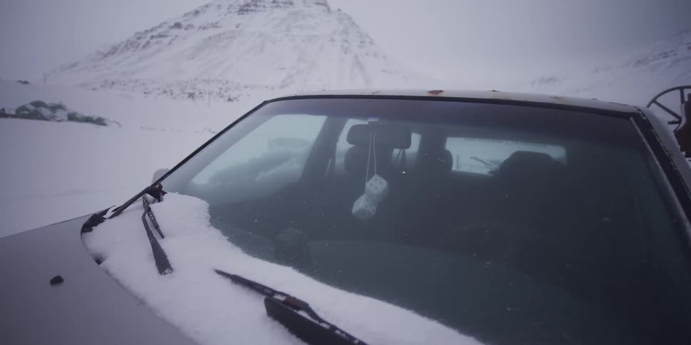 A slow pull-back shot reveals an old, snow-covered car with fuzzy dice hanging from the mirror, abandoned in a desolate, winter mountain landscape