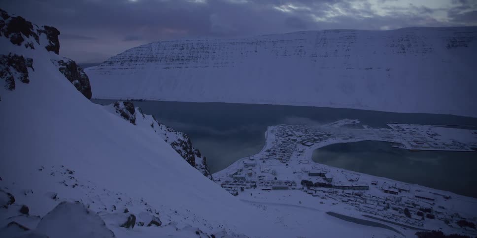 A slow zoom out reveals a remote coastal town nestled between snow-covered mountains and a fjord during a cold winter twilight
