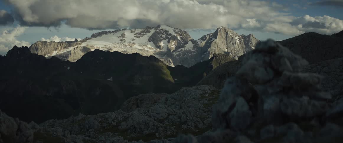 A slow pan across a rocky foreground reveals a majestic, snow-capped mountain range under a dramatic, cloudy sky