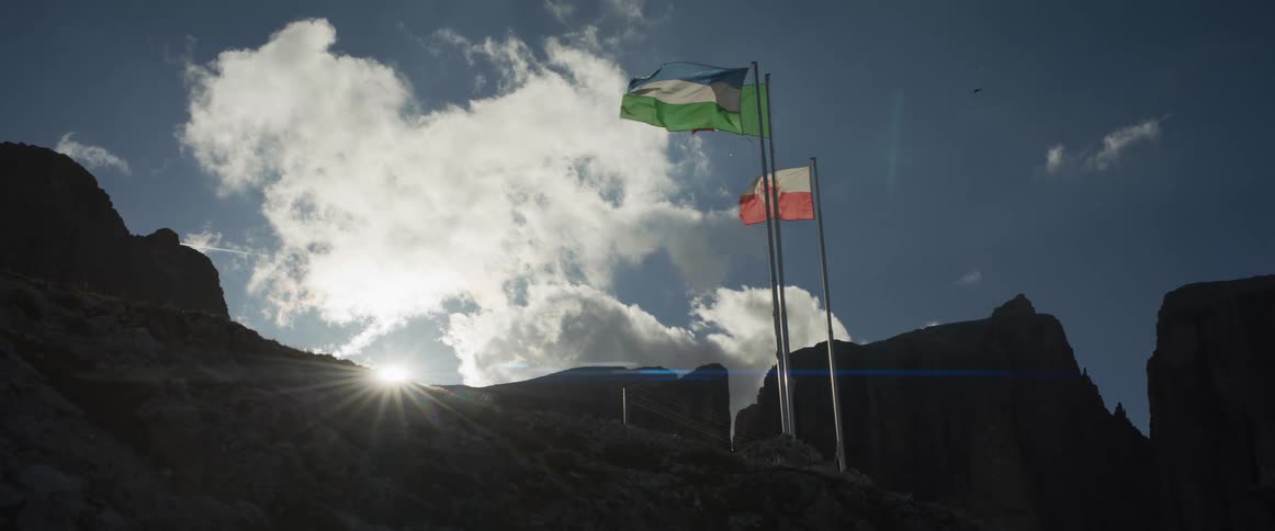 The Ladin and South Tyrol flags wave majestically on a silhouetted mountain summit, backlit by the bright sun and a partly cloudy sky