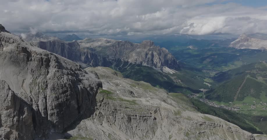 Ascending aerial shot over a vast and rugged mountain range, revealing the breathtaking landscape of peaks and a lush green valley under a cloudy sky