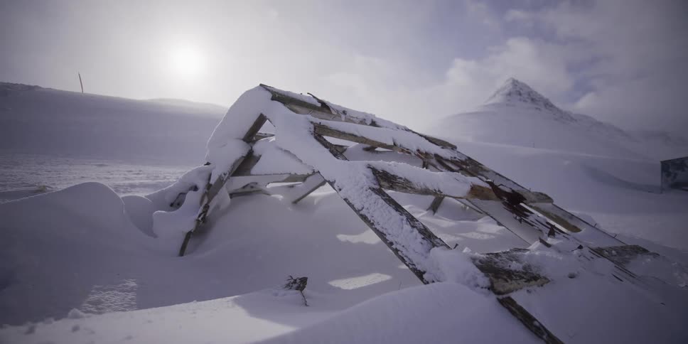 Slow dolly shot past a ruined wooden structure buried in the deep snow of a desolate and windy arctic mountain landscape
