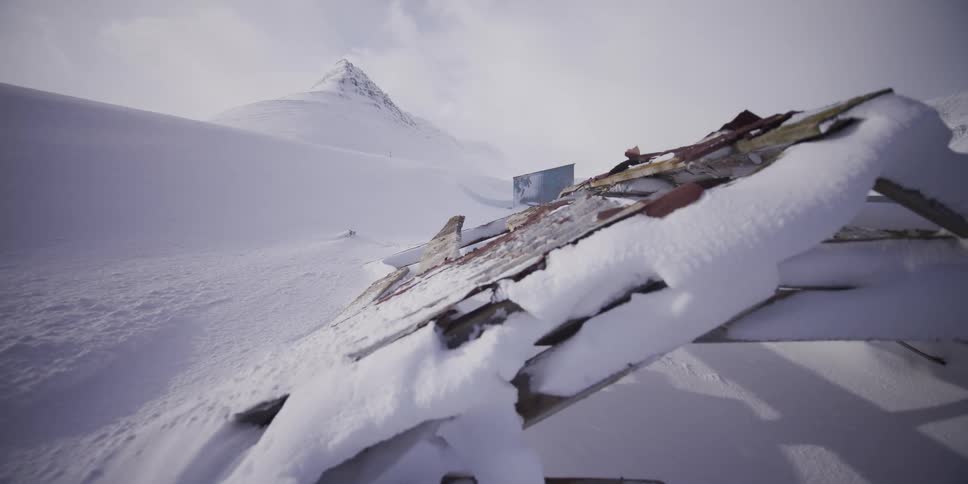 A slow tracking shot reveals the snow-covered remains of a collapsed building in a vast and remote winter mountain landscape