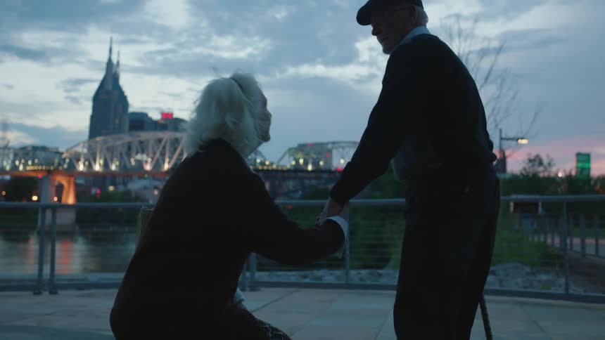 A man stands with the aid of a cane then holds hands with a woman