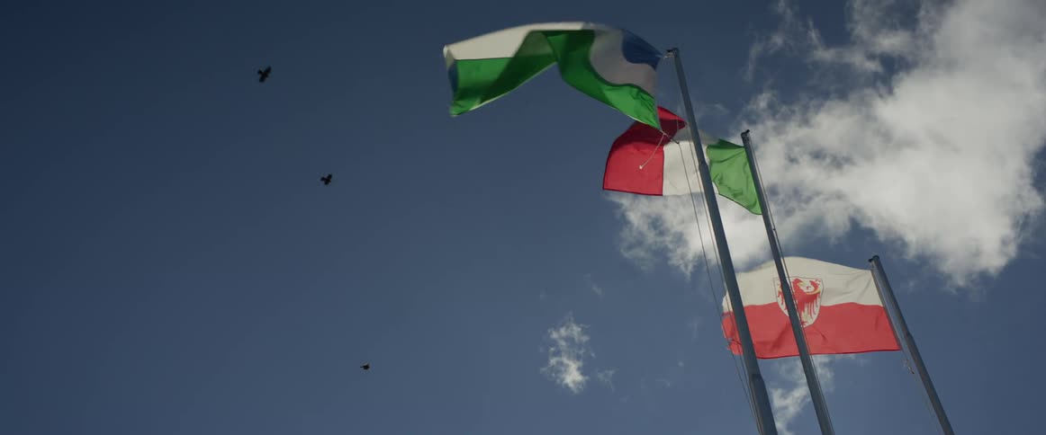 The flags of Ladinia, Italy, and South Tyrol wave gracefully in slow motion against a blue sky as birds fly by