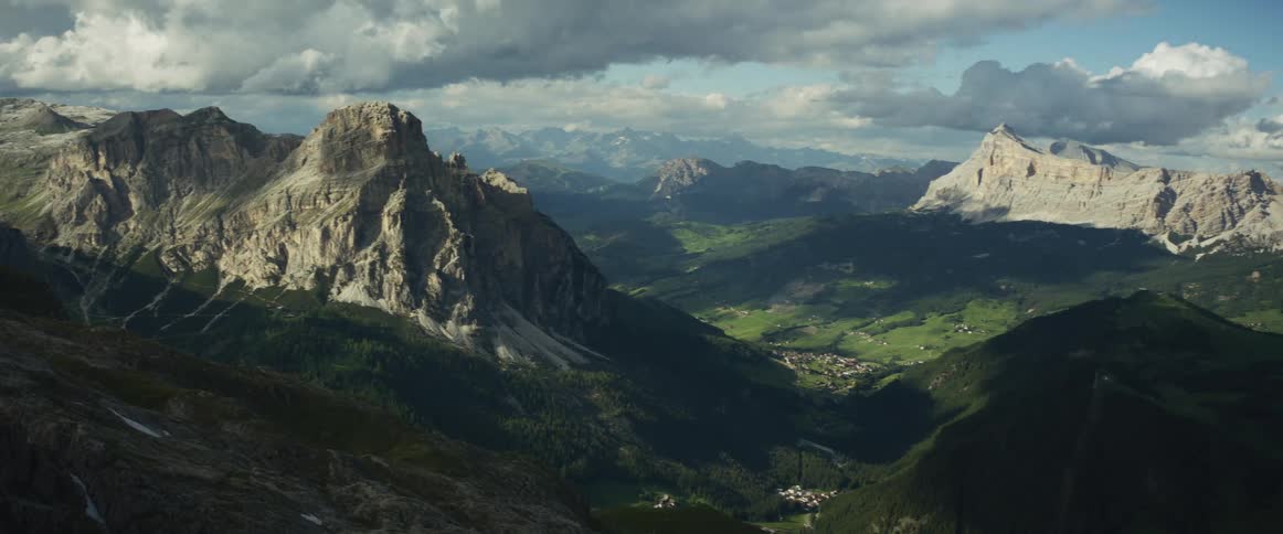 A slow pan reveals a breathtaking view of the rugged Dolomite mountain range and a village nestled in the green valley below