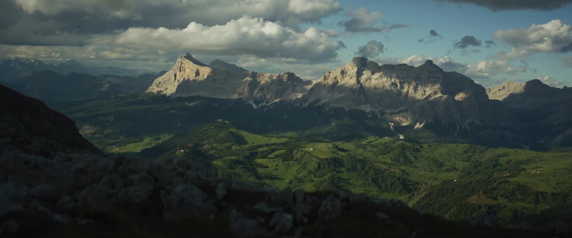 A stunning wide shot reveals the majestic, sun-kissed peaks of a mountain range towering over a lush green valley under a dramatic, cloudy sky