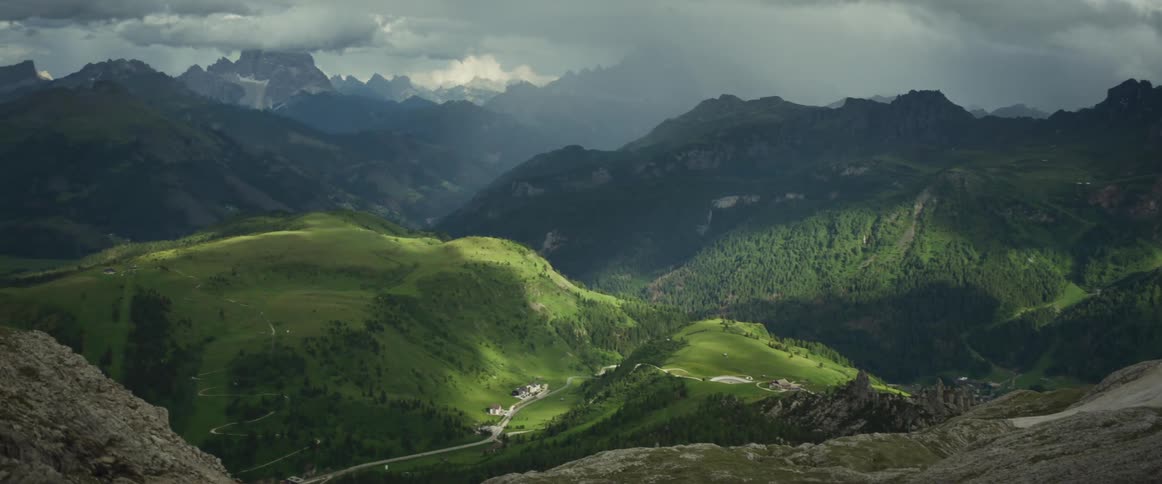 Sunlight breaks through stormy clouds to illuminate a vast, green mountain valley in a slow panning shot