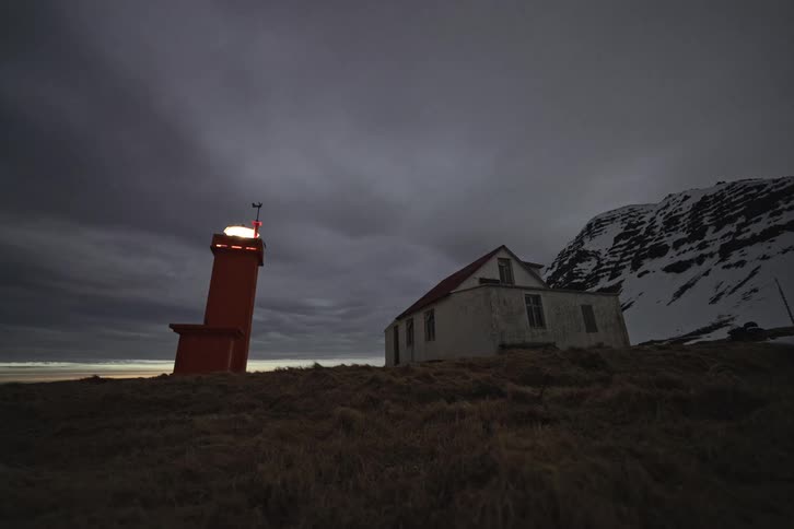 A red lighthouse flashes next to a remote house on a desolate, snowy hillside at twilight