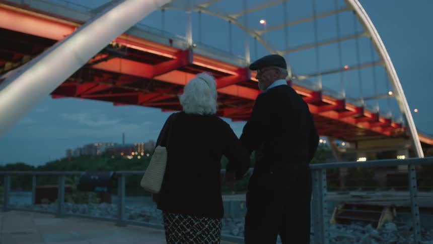 An elderly couple hold hands and walk beneath a city bridge