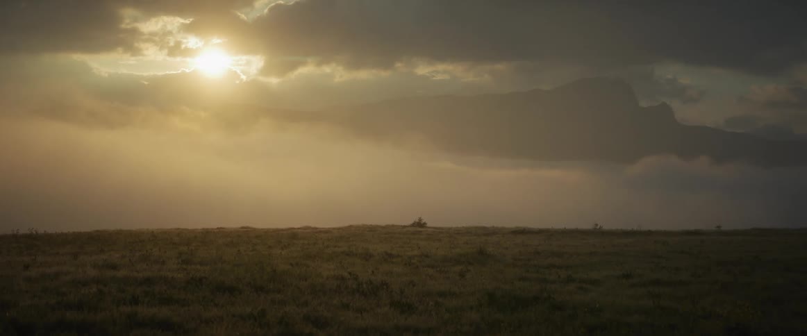 The setting sun casts a warm golden glow through dramatic clouds over a vast, misty mountain landscape