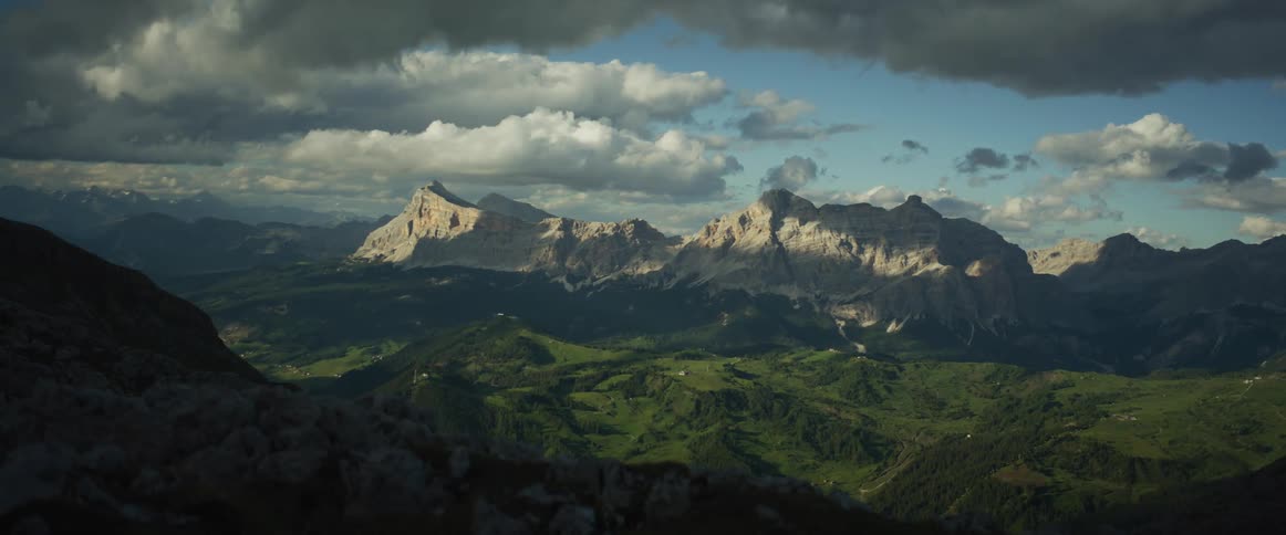 A majestic view of the Dolomite mountains with sunlit peaks and rolling green valleys under a cloudy sky
