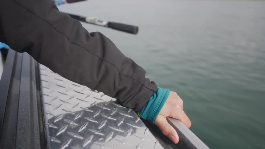 Close-up of a person's hand resting on the diamond plate gunwale of a boat moving through calm water