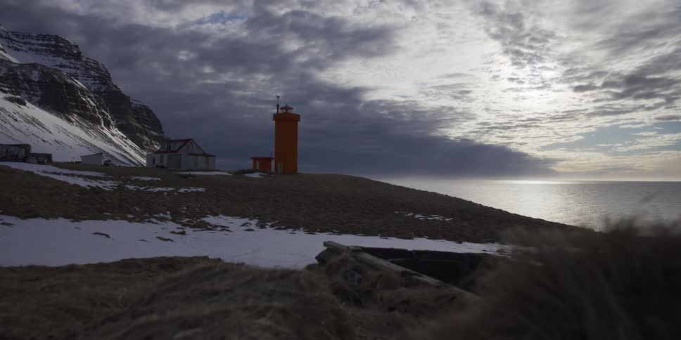A dramatic dolly shot reveals a striking orange lighthouse standing against a snowy mountain and a vast, cloudy ocean view