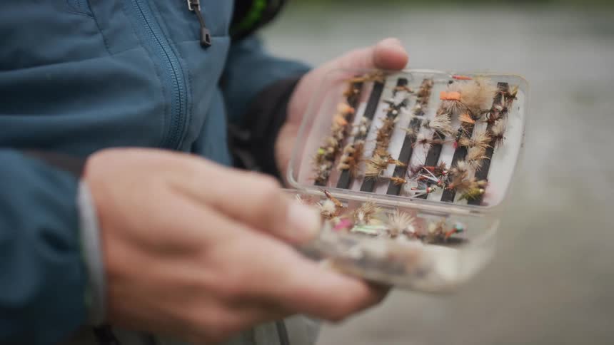 A fly fisherman's hands slowly open a clear plastic tackle box to reveal a collection of colorful handmade flies
