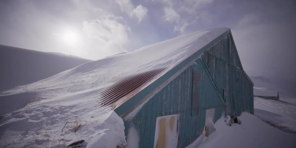 A weathered blue barn is buried in snow drifts as the camera slowly pulls back, revealing a desolate and cold winter scene