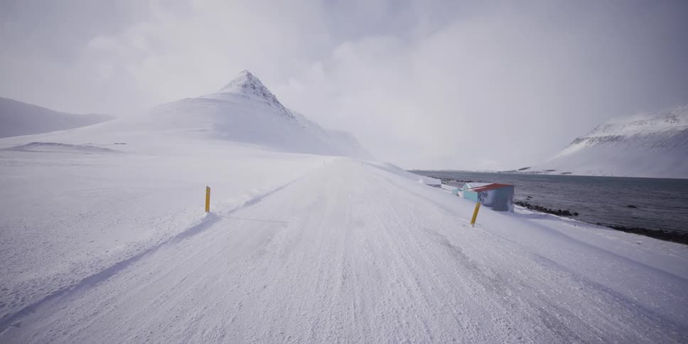 A slow journey down a remote, snow-covered road in a vast, desolate winter landscape with a majestic mountain on the horizon