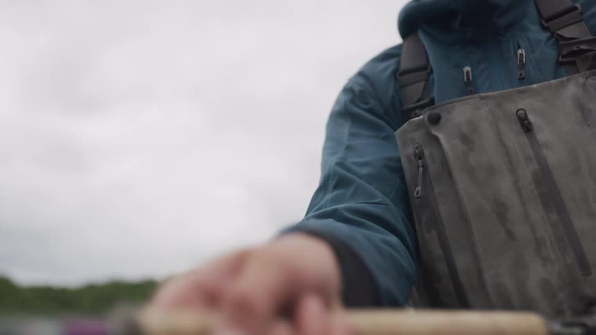 A fly fisherman in waders reels in his fishing line while standing in a river on a cloudy day