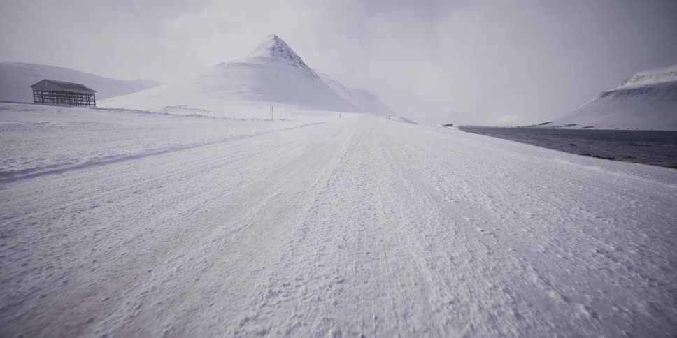 A first-person perspective shot moves along a desolate, snow-covered road as wind blows across the barren arctic landscape