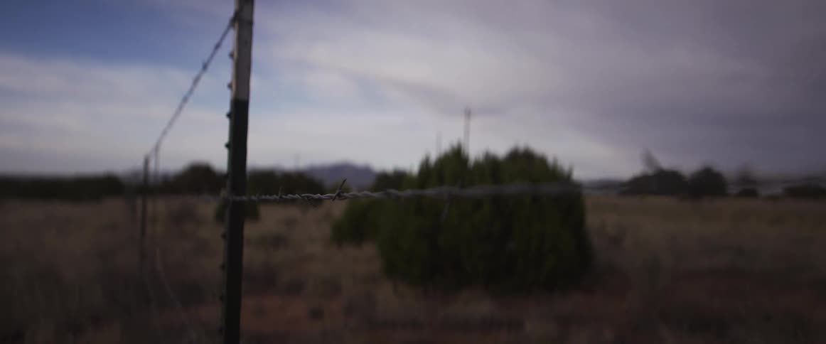 A close-up view of a metal barbed wire fence with an out-of-focus, arid landscape behind it