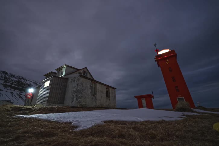 A low-angle time-lapse of a bright orange lighthouse and a weathered house under a cloudy night sky as people with headlamps explore and light paint the scene