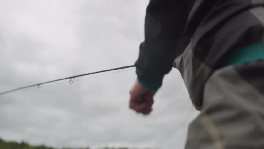 A low-angle, slow-motion shot captures a fly fisherman's hands expertly stripping the fishing line against a cloudy sky