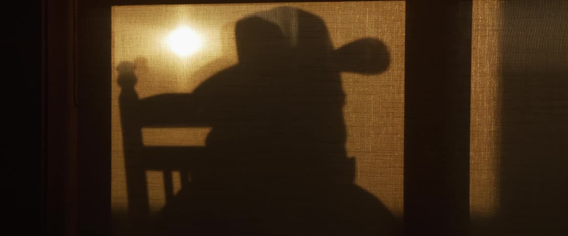 The eerie silhouette of a person in a western hat rocks slowly in a chair behind a warm, glowing curtain