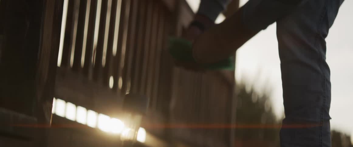 Golden hour sun flares behind a person in slow motion meticulously scrubbing a wooden porch railing