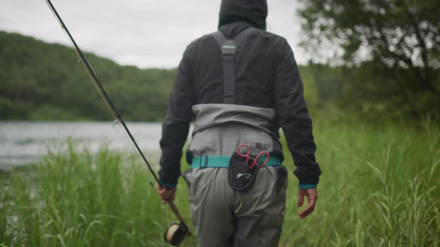 A fly fisherman in full waders and a hooded jacket walks in slow motion through tall grass towards a river