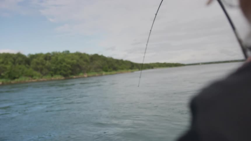 Over-the-shoulder slow-motion shot of a fisherman reeling in a fish with a bent rod on a tranquil river