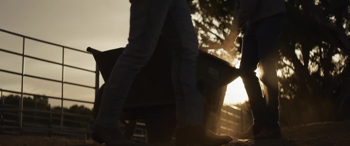 A slow-motion close-up shows two ranchers in boots working together to fill a wheelbarrow on a dusty farm as the sun sets