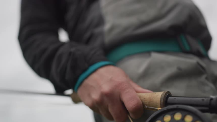 A slow-motion close-up focuses on a fly fisherman's hand holding a rod as the reel is revealed