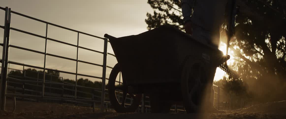 A silhouetted farmer rakes dirt into a wheelbarrow on a dusty ranch as the golden sun sets in the background