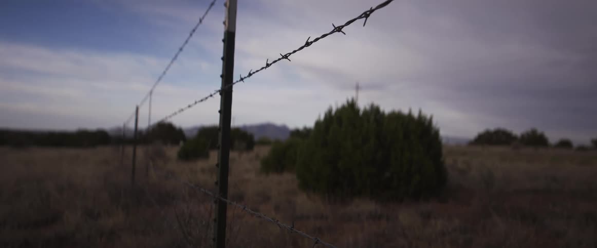 Static close-up of a barbed wire fence dividing a desolate rural landscape under a cloudy sky
