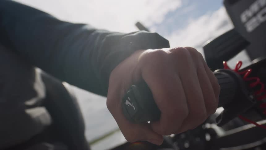 A low-angle, close-up shot shows a hand gripping the tiller handle of a boat motor under a bright, sunny sky