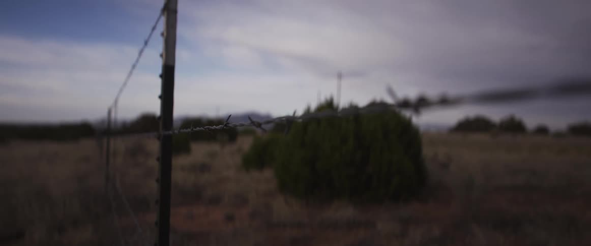 A slow rack focus travels along a strand of barbed wire in a desolate rural landscape under a cloudy sky
