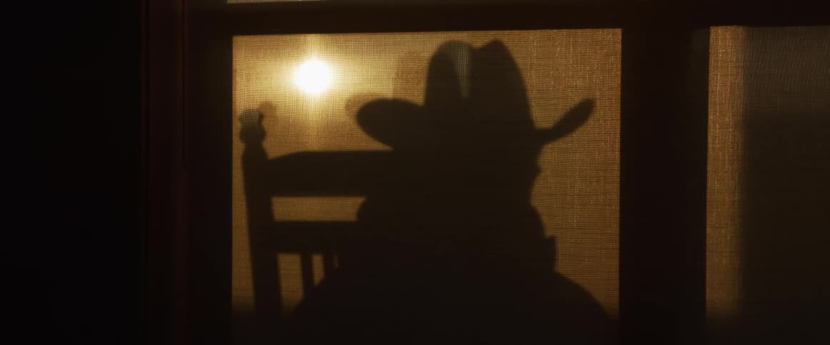 A mysterious silhouette of a cowboy in a rocking chair is cast onto a backlit window screen